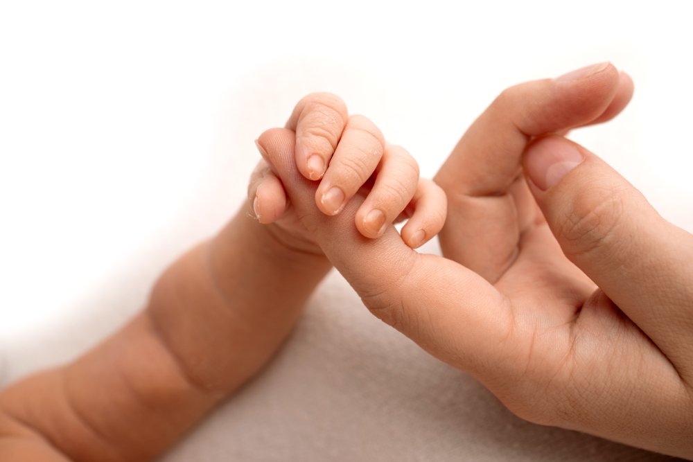 The newborn baby in a white closes has a firm grip on the parent's finger after birth. Close-up little hand of child and palm of mother and father. Parenting, childcare and healthcare concept